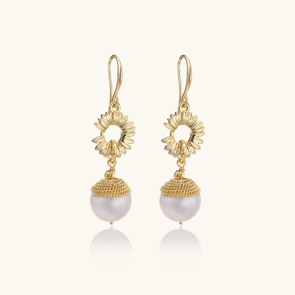 Gold sunburst charm earrings with baroque pearl drops on French hooks against cream background
